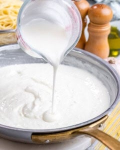 Thick cottage cheese Alfredo sauce being poured from a measuring cup into a skillet with sautéed garlic.
