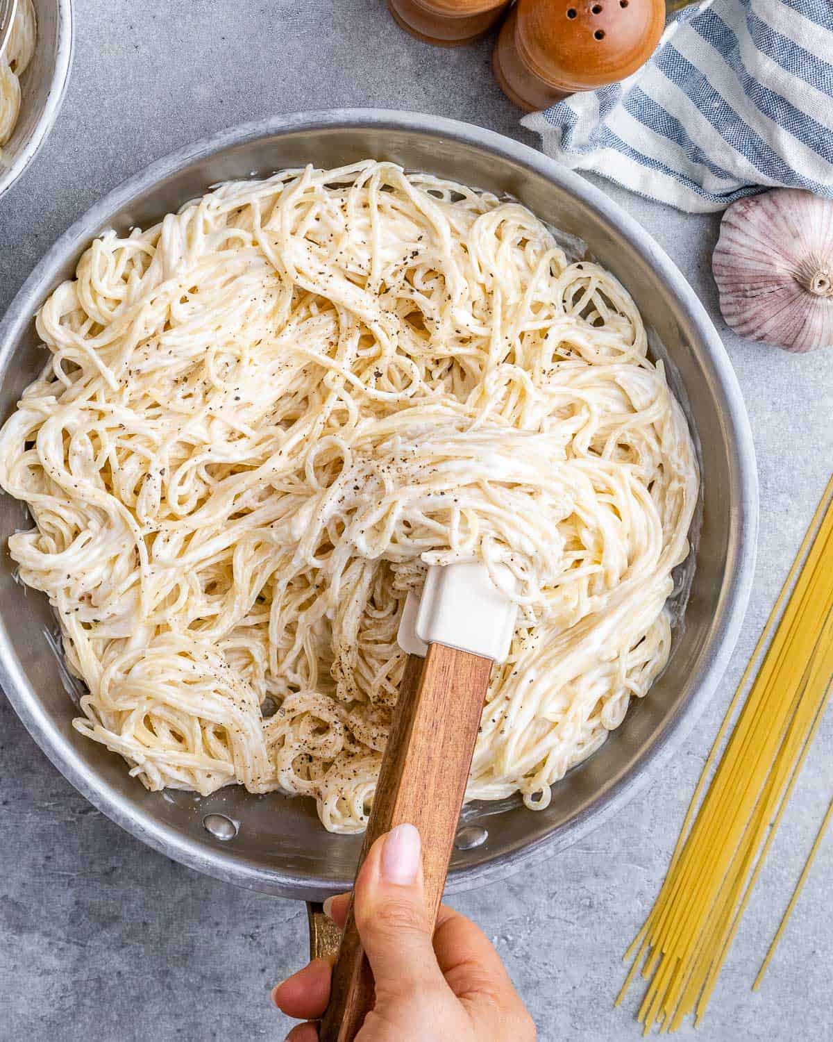 Hand stirring cooked spaghetti in a pan to evenly combine with the Alfredo sauce.
