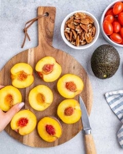 Halved peaches with pits removed on a cutting board, next to cherry tomatoes, pecans, and an avocado.