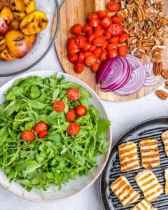 Arugula and cherry tomatoes in a bowl beside sliced red onions, toasted pecans, grilled peaches, and halloumi on a cutting board.