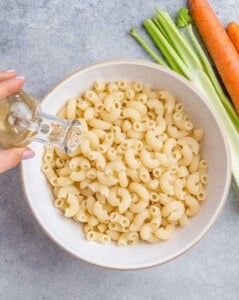 Apple cider vinegar being poured over freshly cooked elbow macaroni in a white bowl.