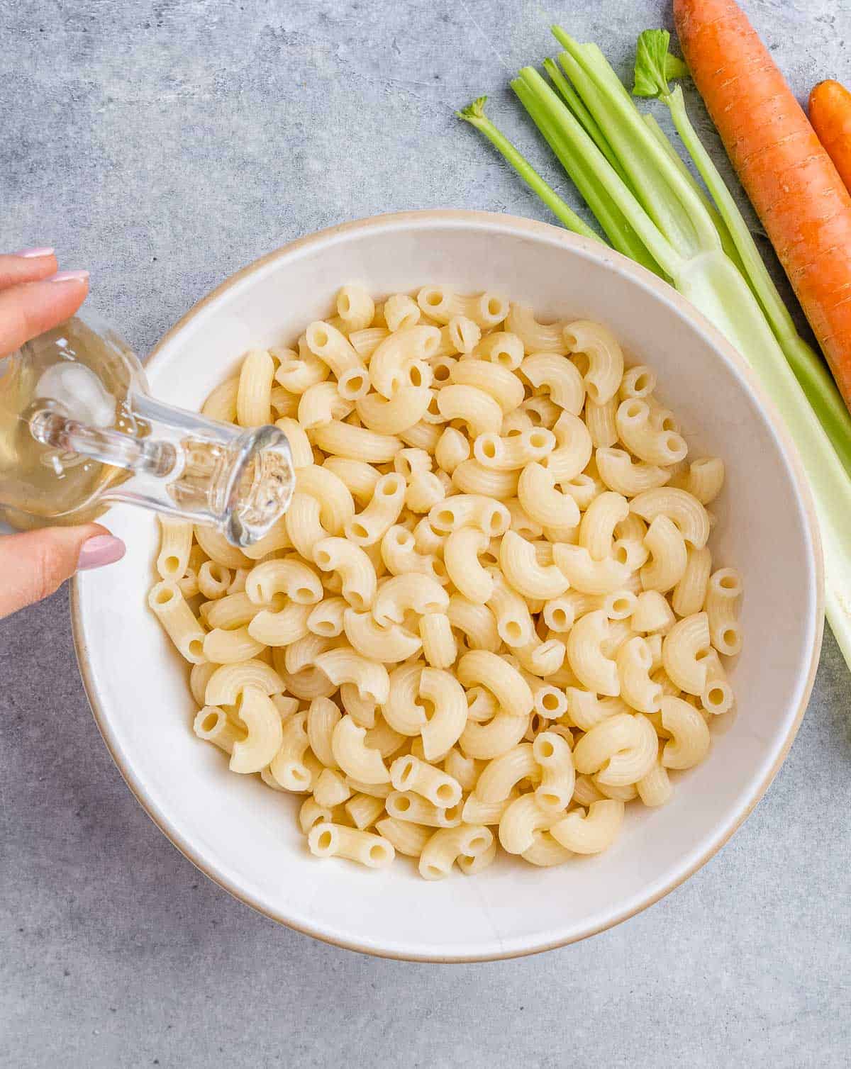 Apple cider vinegar being poured over freshly cooked elbow macaroni in a white bowl.