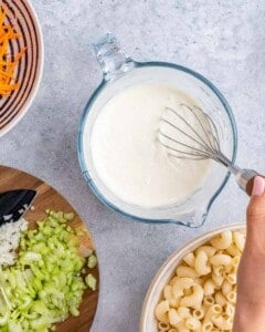 Hand whisking together mayonnaise and milk in a glass measuring cup, with chopped celery, onion, and cooked macaroni nearby.