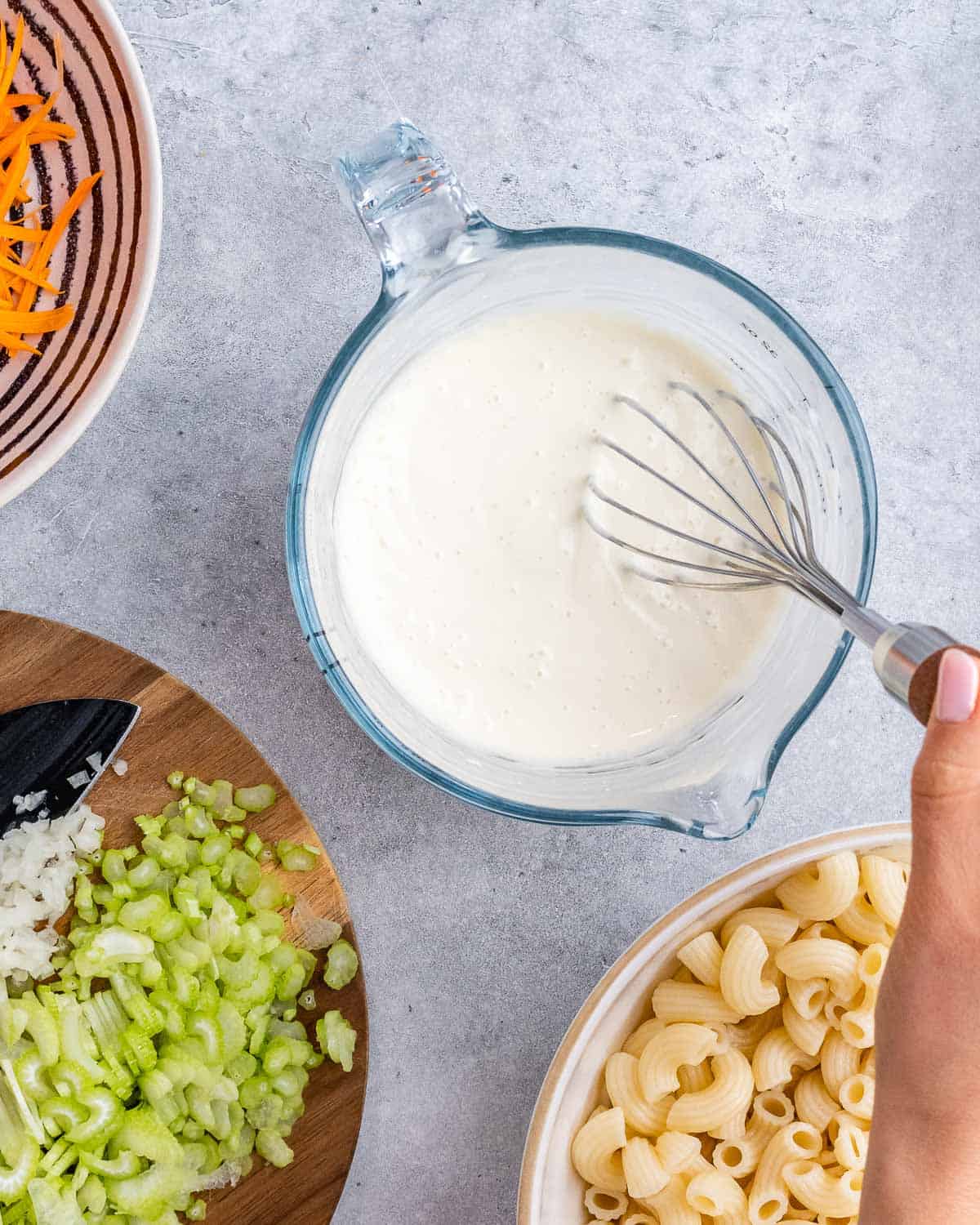 Hand whisking together mayonnaise and milk in a glass measuring cup, with chopped celery, onion, and cooked macaroni nearby.