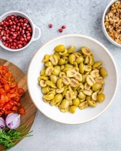 Bowl of halved green olives surrounded by prepped salad ingredients like tomatoes, parsley, and onion.