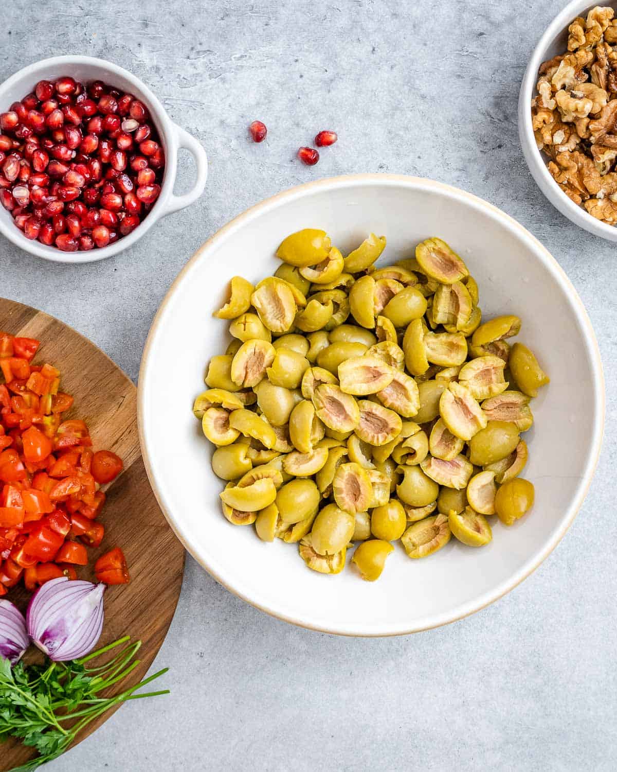 Bowl of halved green olives surrounded by prepped salad ingredients like tomatoes, parsley, and onion.