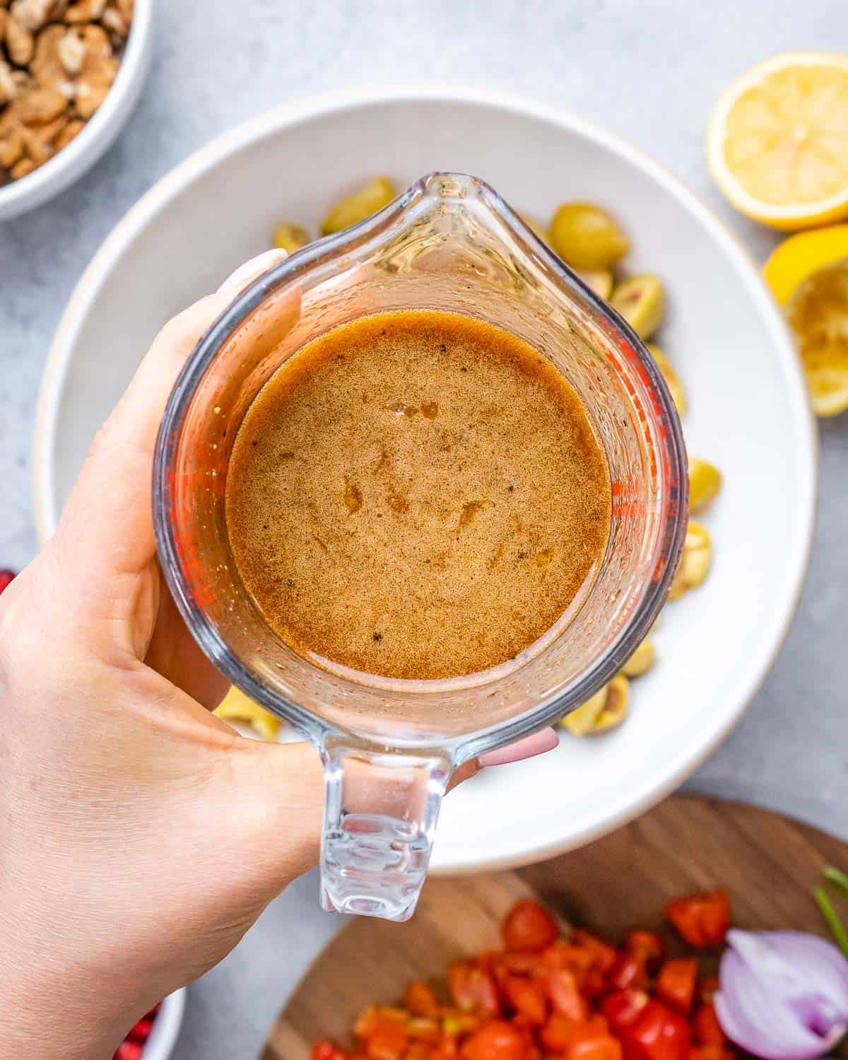 Hand holding a glass measuring cup filled with smoky pomegranate molasses dressing.
