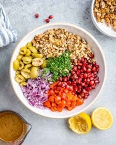 Large mixing bowl with olives, walnuts, parsley, tomatoes, pomegranate seeds, and red onion arranged neatly.
