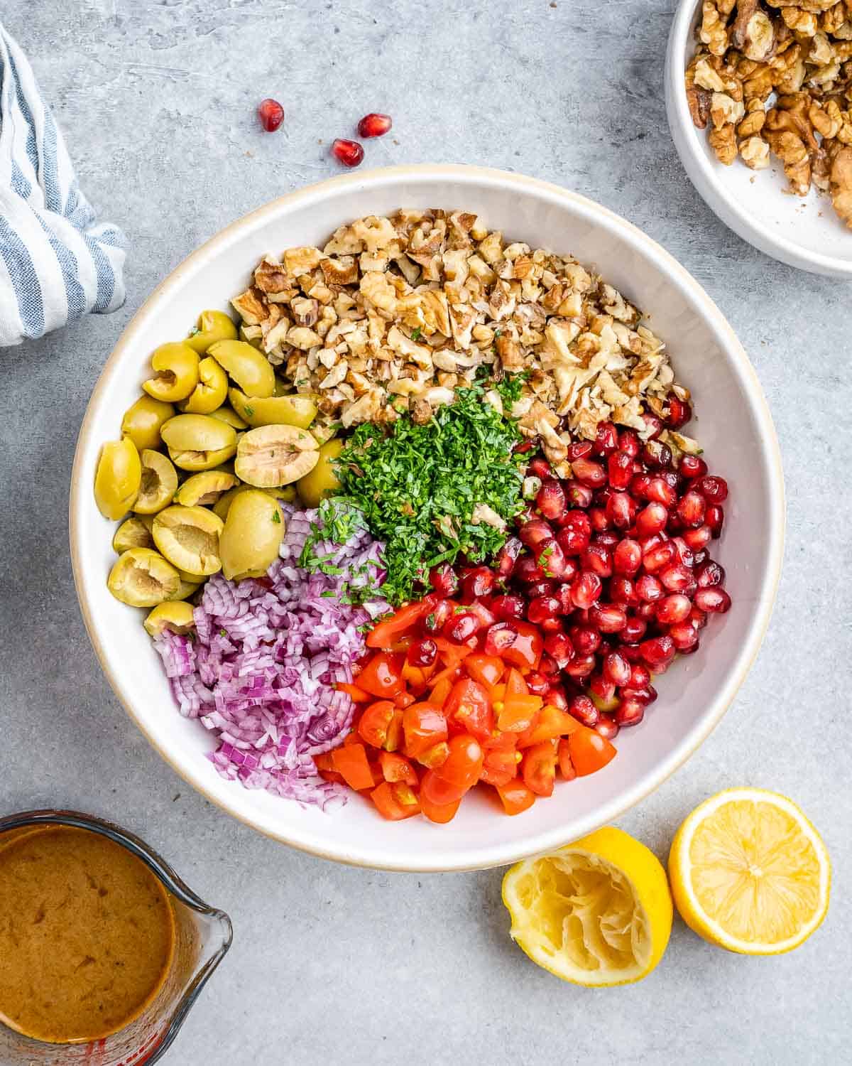 Large mixing bowl with olives, walnuts, parsley, tomatoes, pomegranate seeds, and red onion arranged neatly.