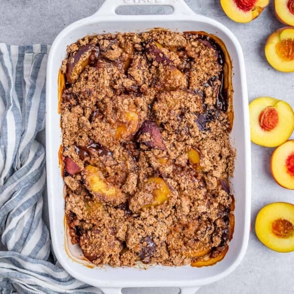 A white baking dish with baked peach cobbler and fresh sliced peaches on the right side of the dish.