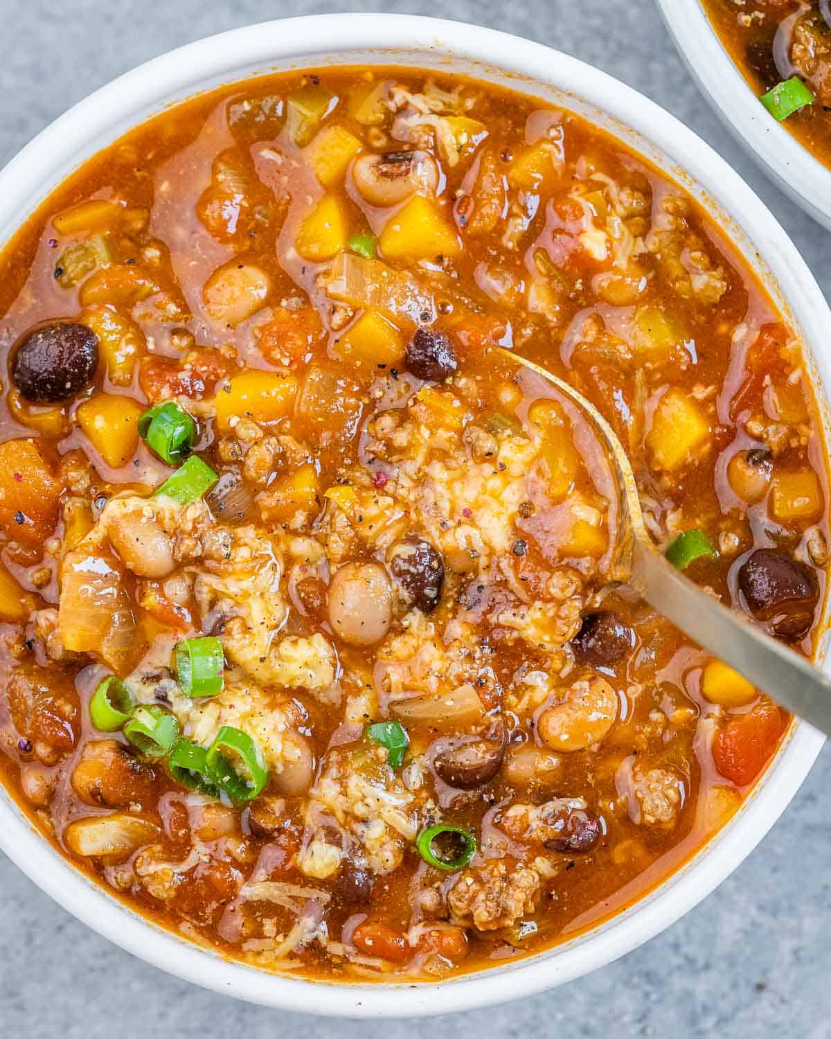 Spoon in a bowl full of beef and bean chili with sliced green onion garnishes.