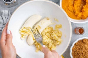 Mashing bananas with a fork in a white bowl.