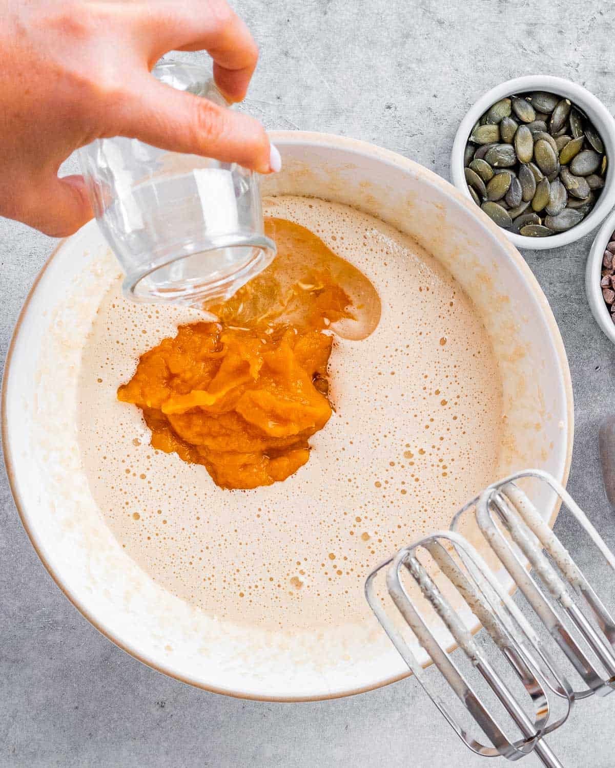 Adding pumpkin puree and oil into egg and sugar mixture in a white bowl.