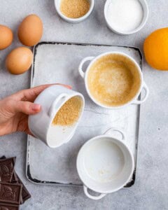 Hand coating the inside of a white ramekin with brown sugar after spraying it with oil, preparing it for baking.
