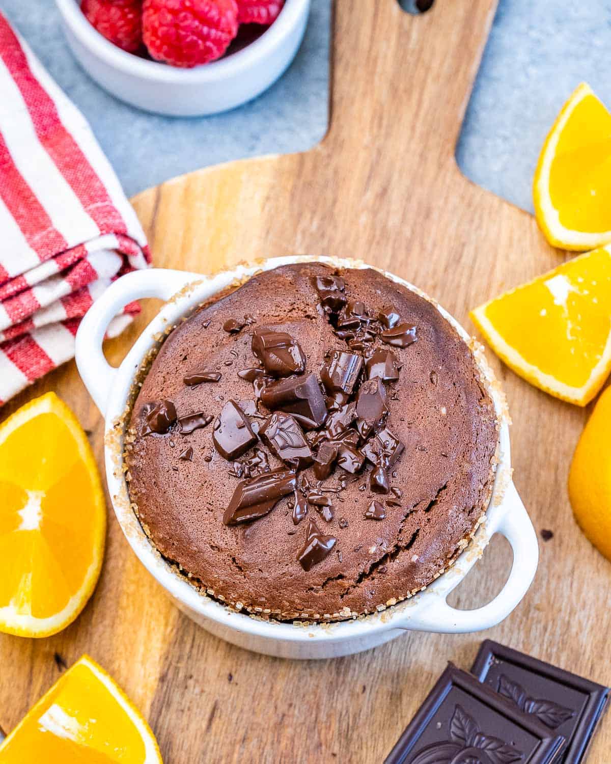 Freshly baked Chocolate Soufflé in a white ramekin, topped with chopped chocolate pieces and surrounded by orange slices.