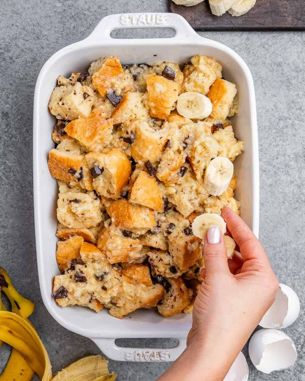 bread pudding mixing added to a baking dish, and a hand adding sliced bananas over it.