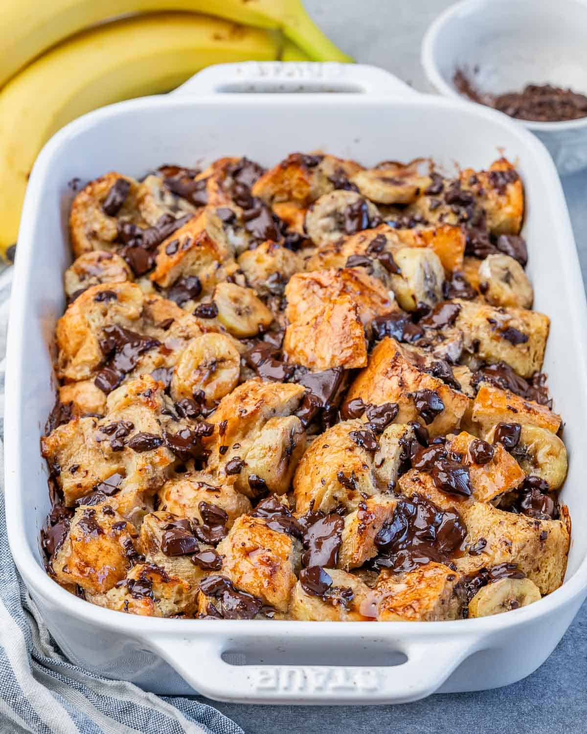 a white baking dish with baked banana bread pudding topped with chocolate and sliced banana.