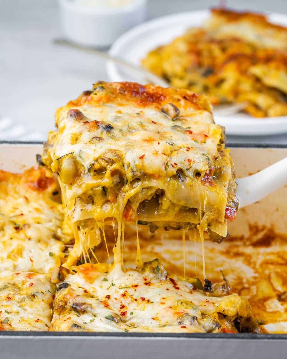 A spatula lifting a gooey, cheesy piece of butternut squash lasagna from a baking dish with stretchy melted cheese pulling away.