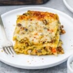 A plated slice of butternut squash lasagna topped with melted mozzarella and red pepper flakes, served with a fork on a white plate.
