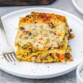 A plated slice of butternut squash lasagna topped with melted mozzarella and red pepper flakes, served with a fork on a white plate.