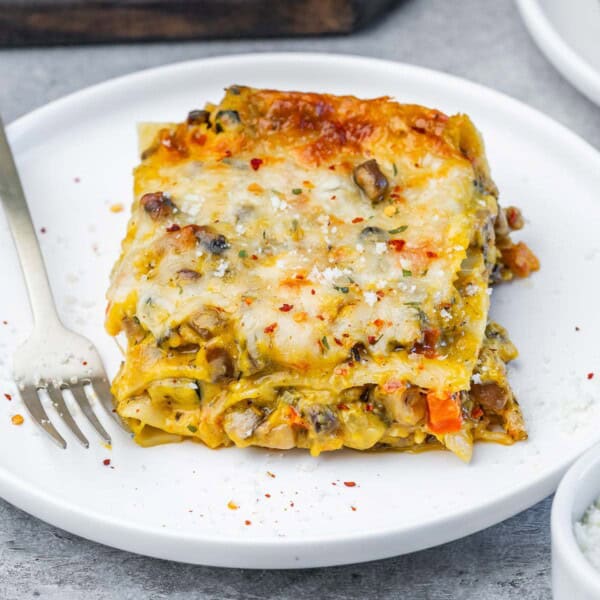 A plated slice of butternut squash lasagna topped with melted mozzarella and red pepper flakes, served with a fork on a white plate.