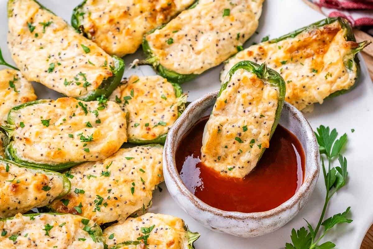 A plate with jalapeno poppers and a dipping sauce bowl with one stuffed jalapeno in the bowl.