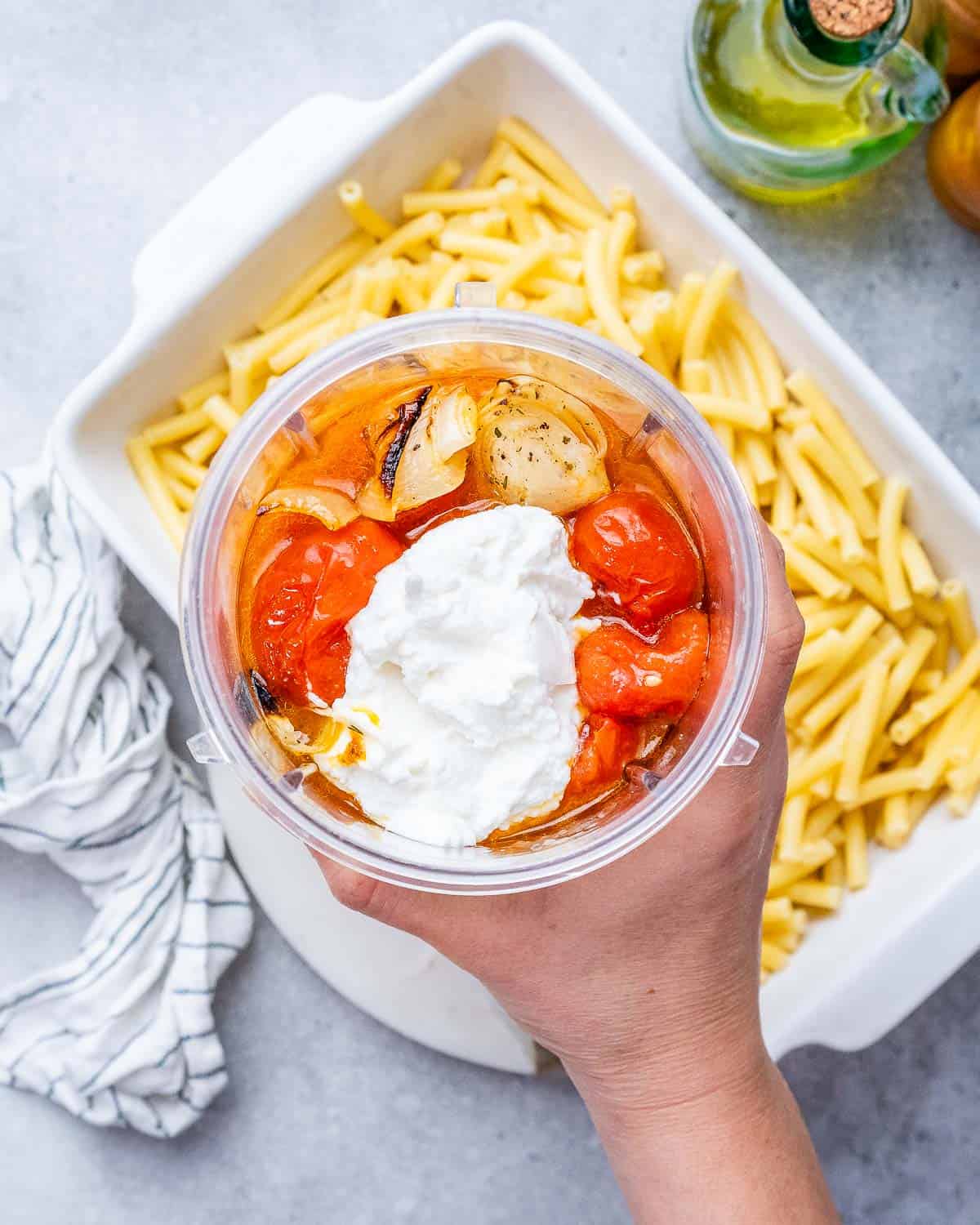 Hand holding a plastic container over pasta in a casserole dish.