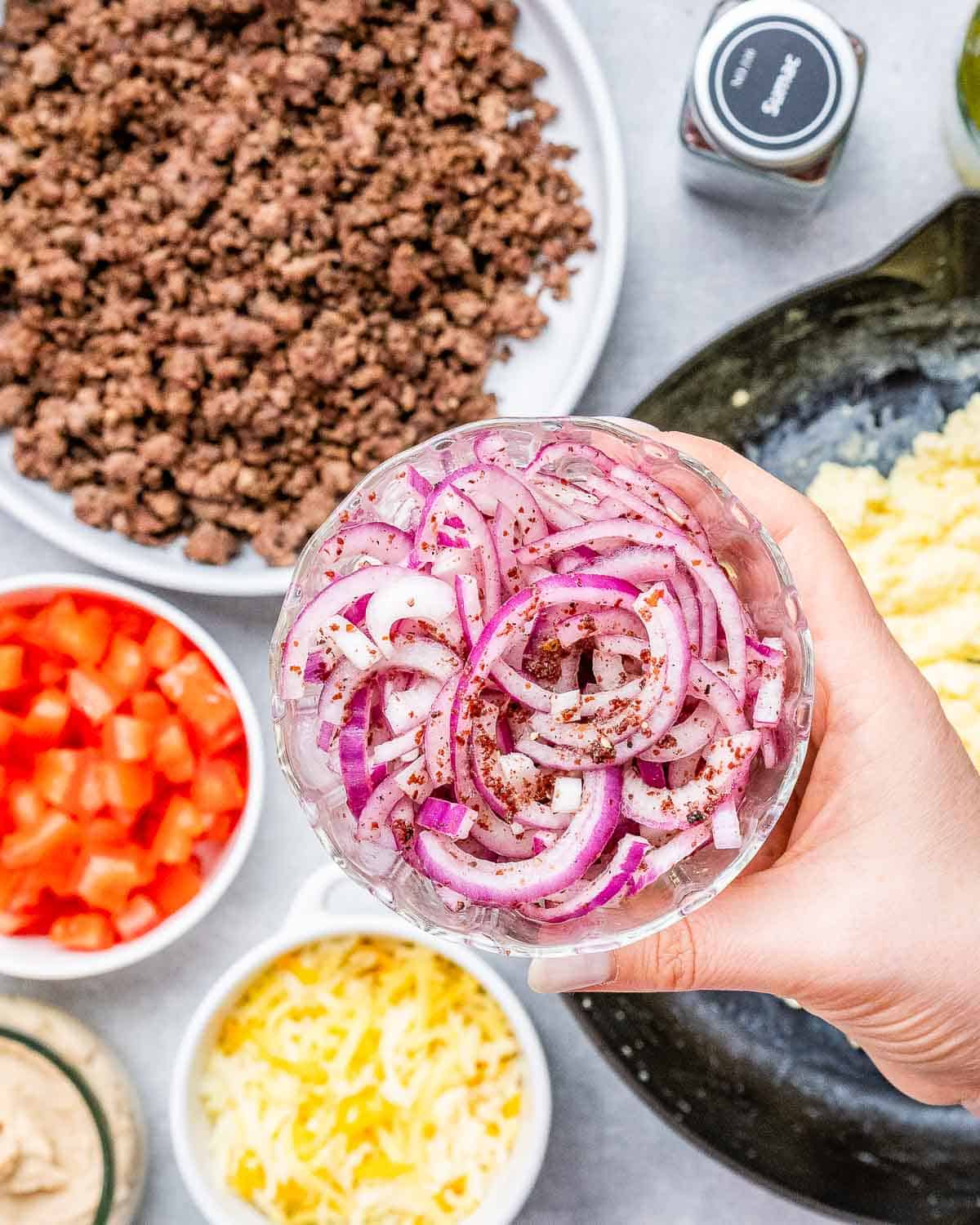 A hand holding a bowl of sliced onions over the ingredients to make burritos.