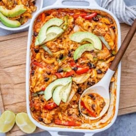 a spoon in a baking dish with chicken fajita, topped with sliced avocado.