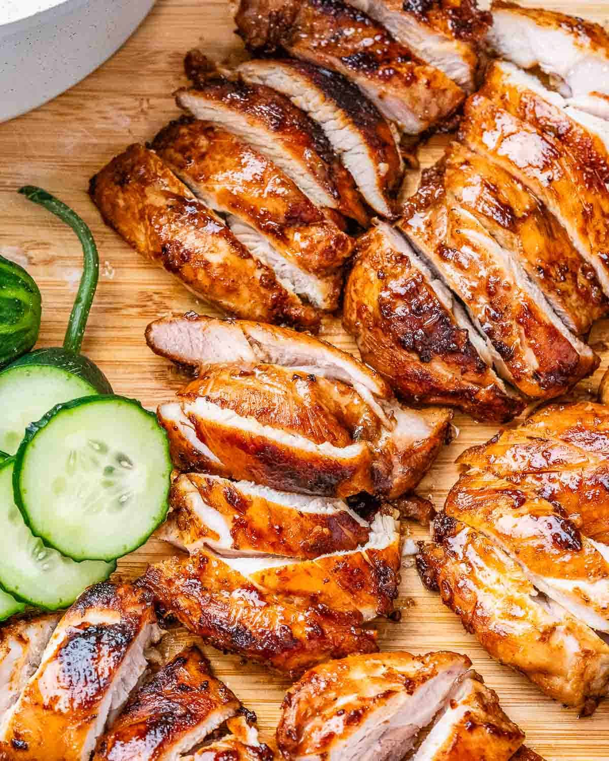 sliced cooked chicken thighs on a cutting board with sliced cucumbers.