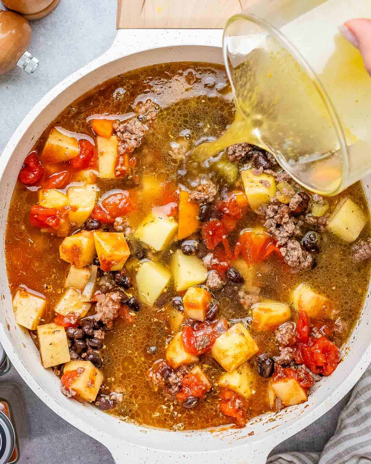 pouring beef broth into round pot of cowboy soup with vegetables and ground beef