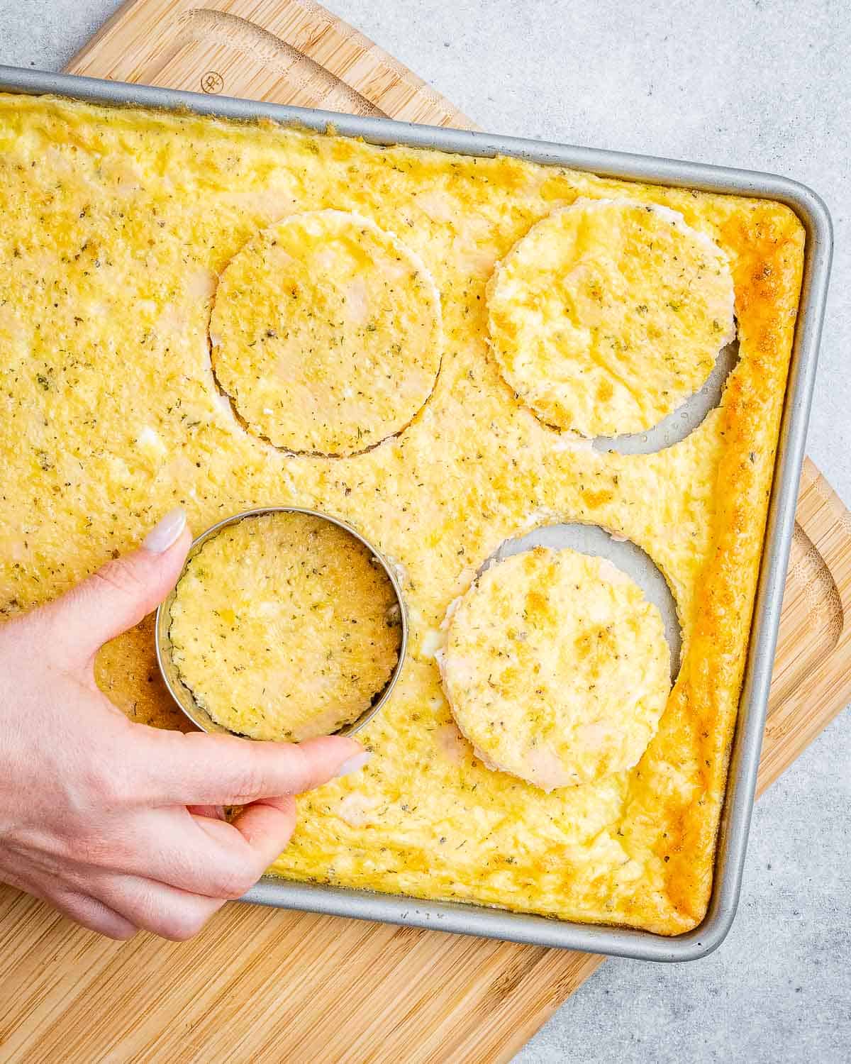 A hand cutting out circles from a sheet pan of cooked eggs.