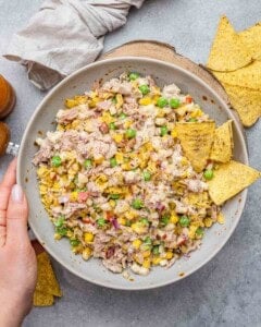 Hands holding a bowl of finished tuna salad with visible peas, corn, red onion, and chips.