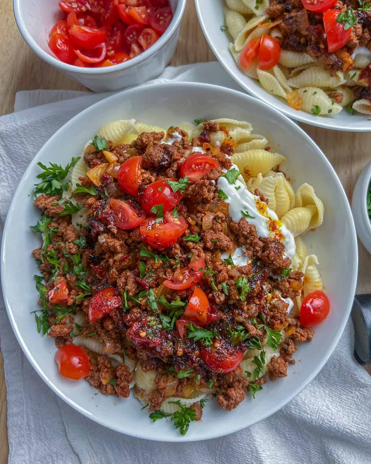 A white dish with pasts topped with seasoned beef and garnished with chopped tomatoes and parsley.