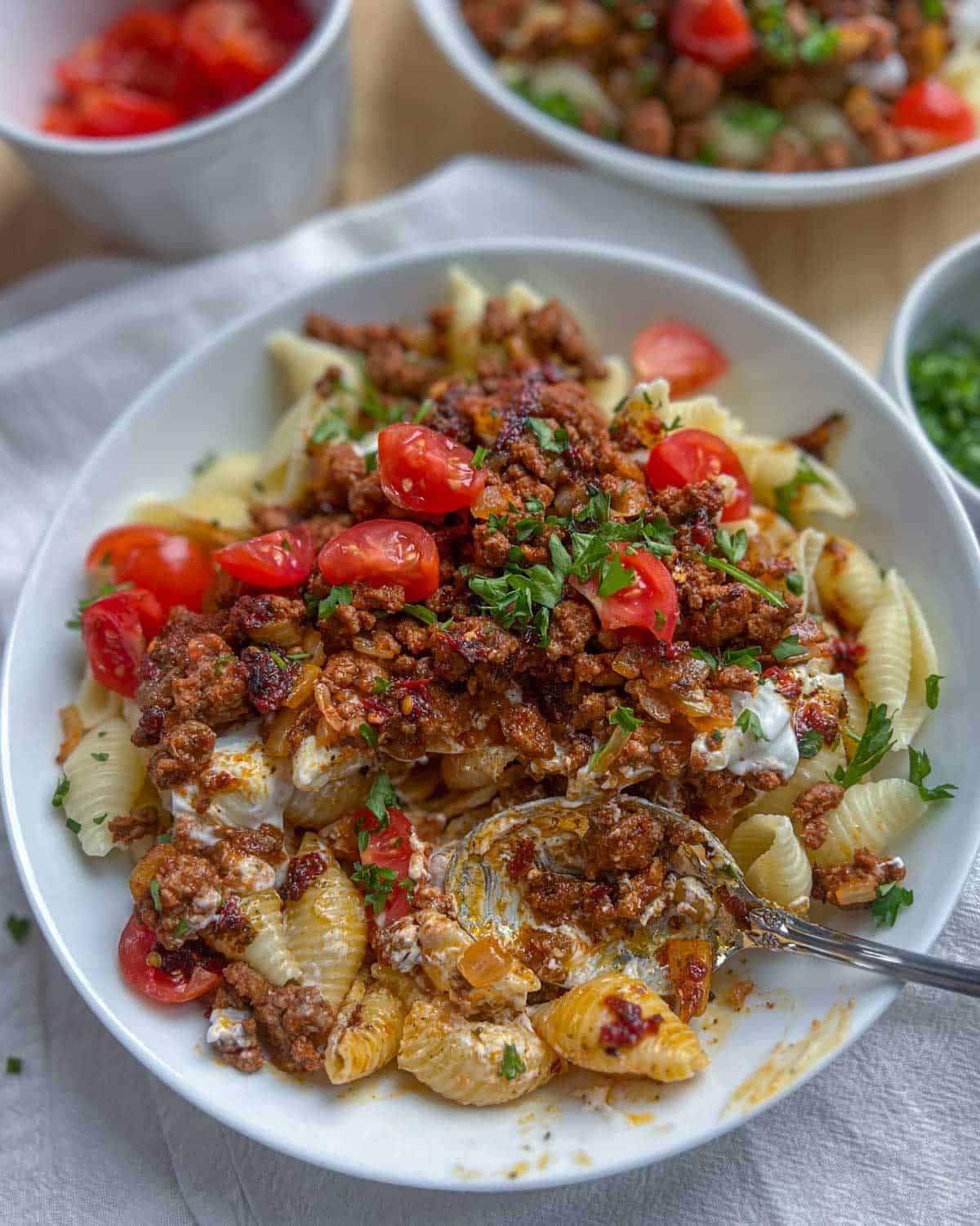 A spoon in a pasta dish with meat sauce topped with chopped tomatoes and parsley.