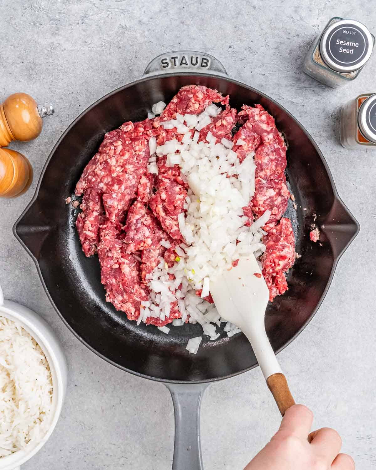 A white spatula stirring onions and raw ground beef in a skillet.