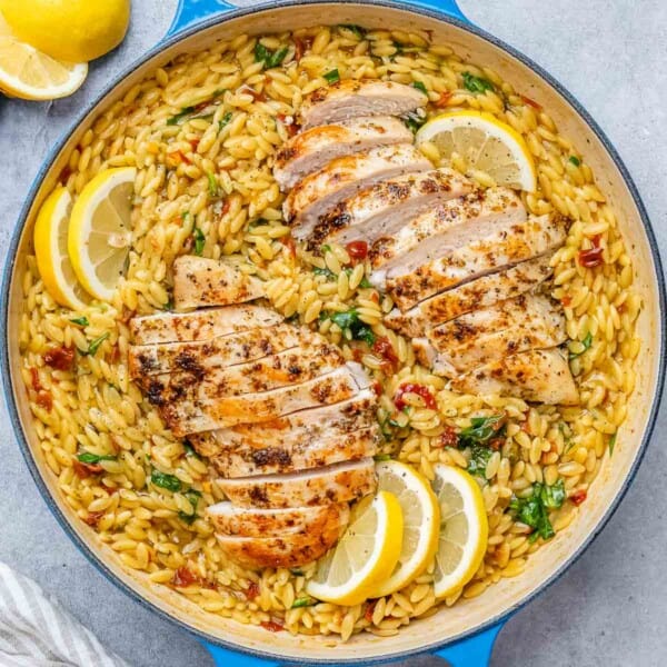 A blue skillet with creamy orzo, topped with sliced chicken breast and lemon wedges.