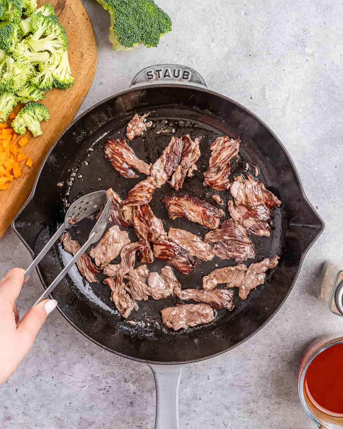 Tongs flipping pieces of meat in a skillet.