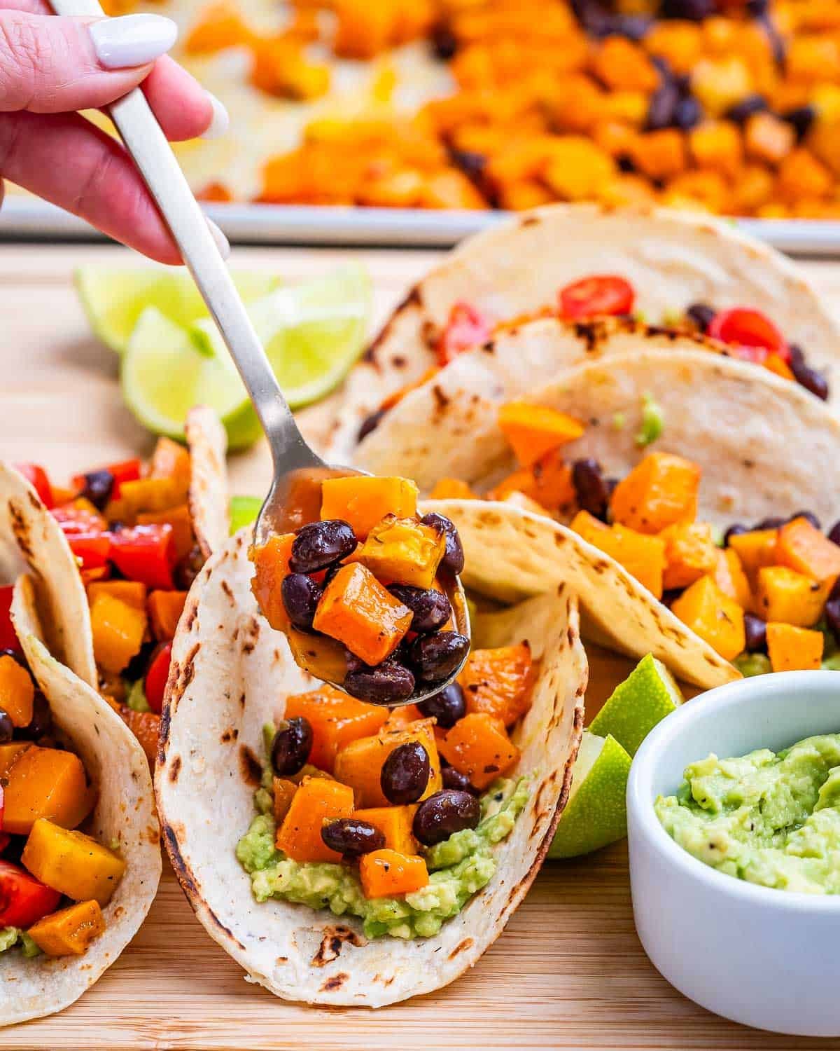 Close up view of tacos being assembled, with a spoon adding filling into a tortilla, surrounded by guacamole and lime.
