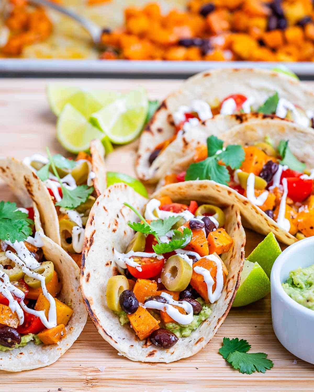 Assembled tacos with a colorful mix of sweet potato, black beans, tomatoes, and olives, topped with cilantro and lime wedges on the side.