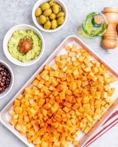 Diced sweet potatoes tossed with olive oil and seasoning, ready to be roasted, on a sheet pan next to other taco ingredients.