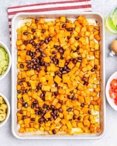 Roasted diced sweet potatoes and black beans spread on a baking sheet, surrounded by small bowls of ingredients.