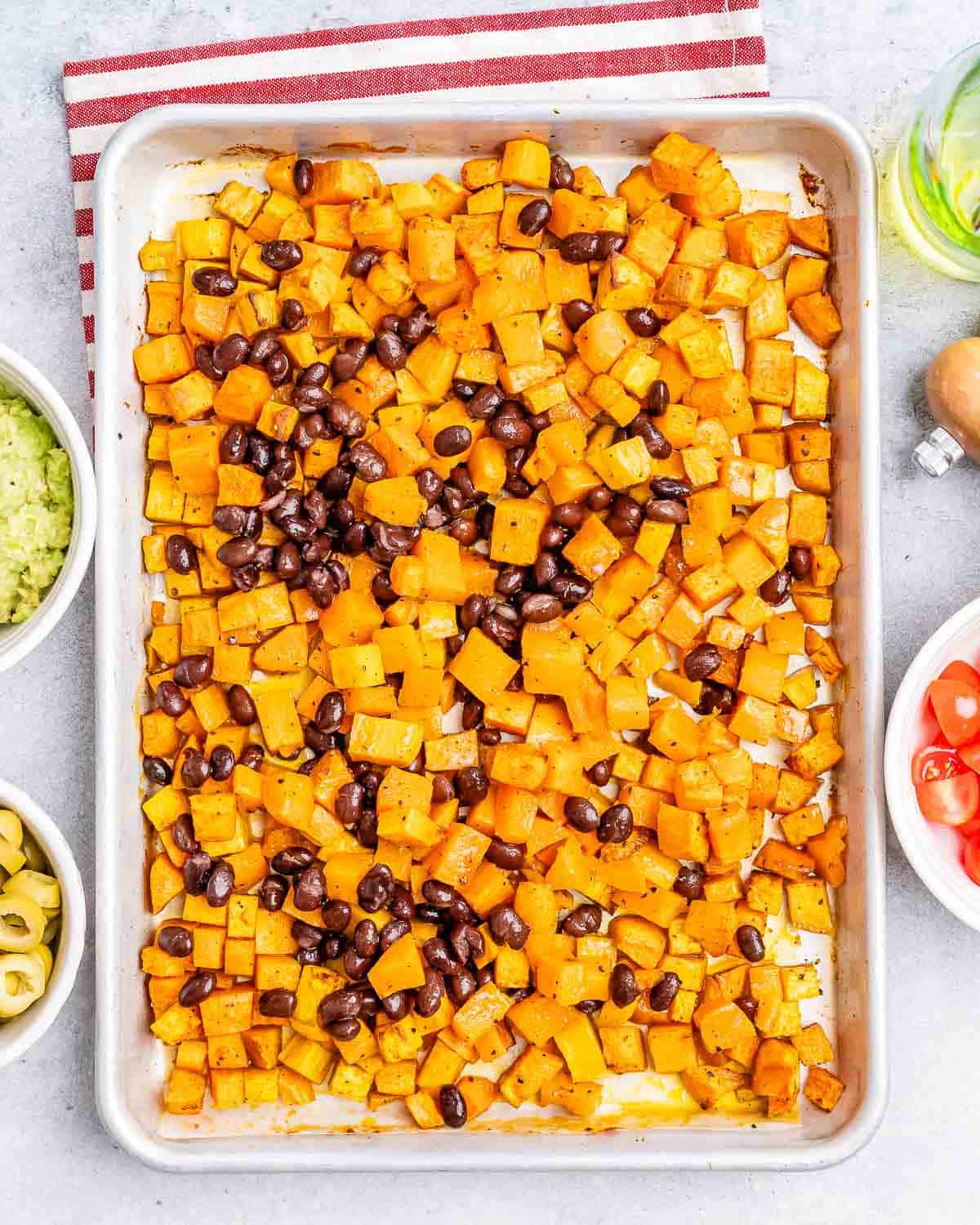 Roasted diced sweet potatoes and black beans spread on a baking sheet, surrounded by small bowls of ingredients.