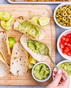 Tortillas being filled with guacamole on a wooden board, surrounded by small bowls of tomatoes, olives, and lime wedges.