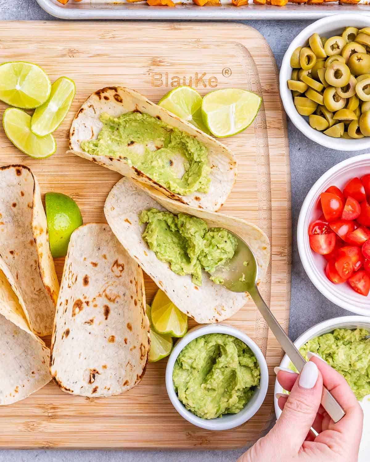 Tortillas being filled with guacamole on a wooden board, surrounded by small bowls of tomatoes, olives, and lime wedges.