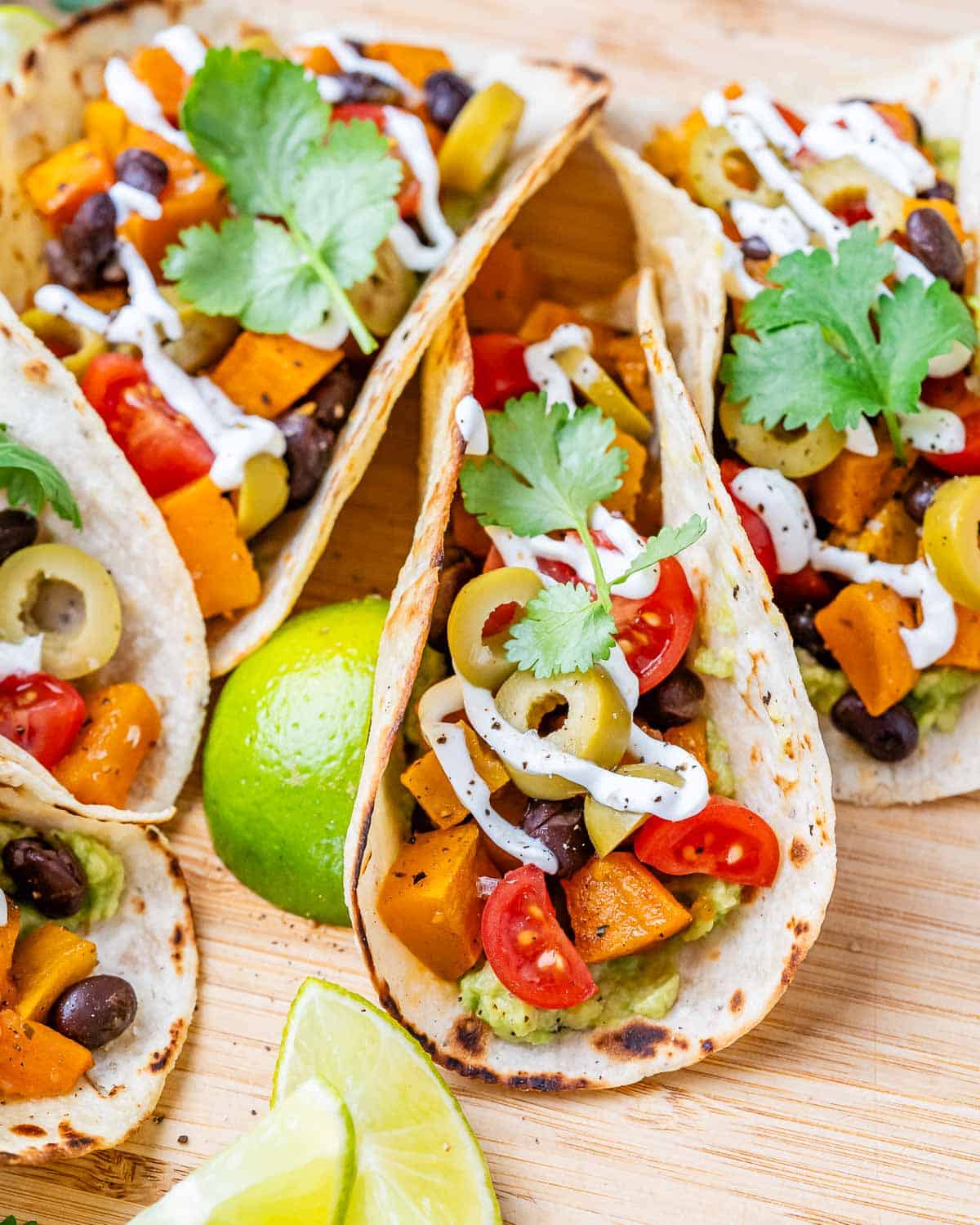A few assembled sweet potato and black bean tacos topped with cherry tomatoes, olives, and cilantro, served with lime wedges and guacamole.