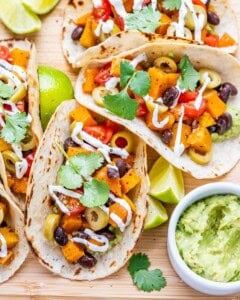 Three tacos on a board filled with sweet potato, black bean, and veggie toppings, served with guacamole and lime wedges.