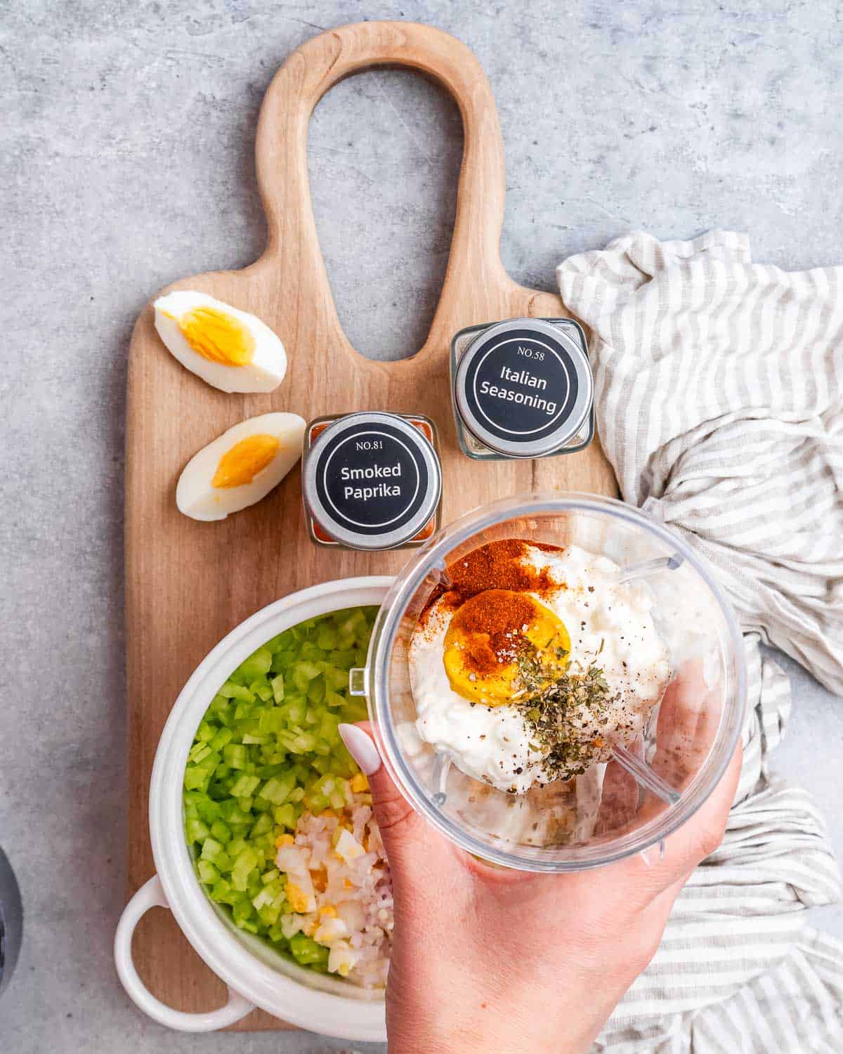 A hand holding a glass bowl with seasonings over a cutting board and a bowl of ingredients to make egg salad.