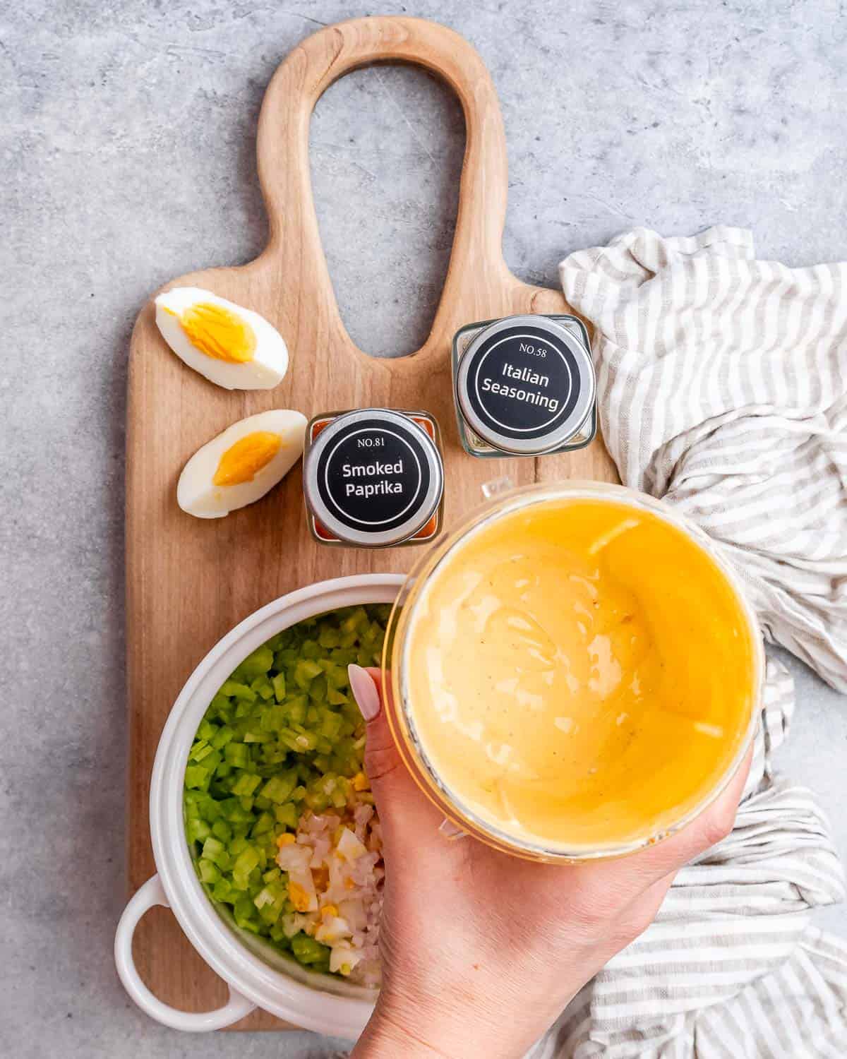 A hand holding a cup with liquid over a cutting board with ingredients to make egg salad.