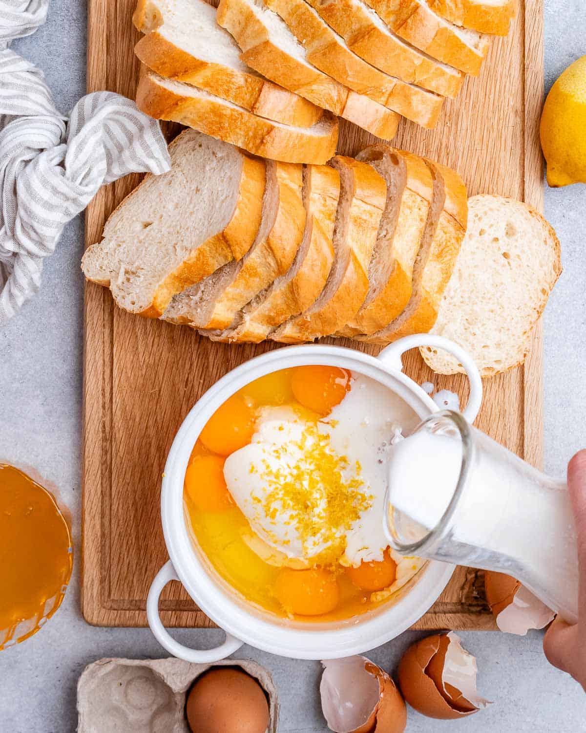 Pouring milk into a dish with eggs with sliced bread on a cutting board.